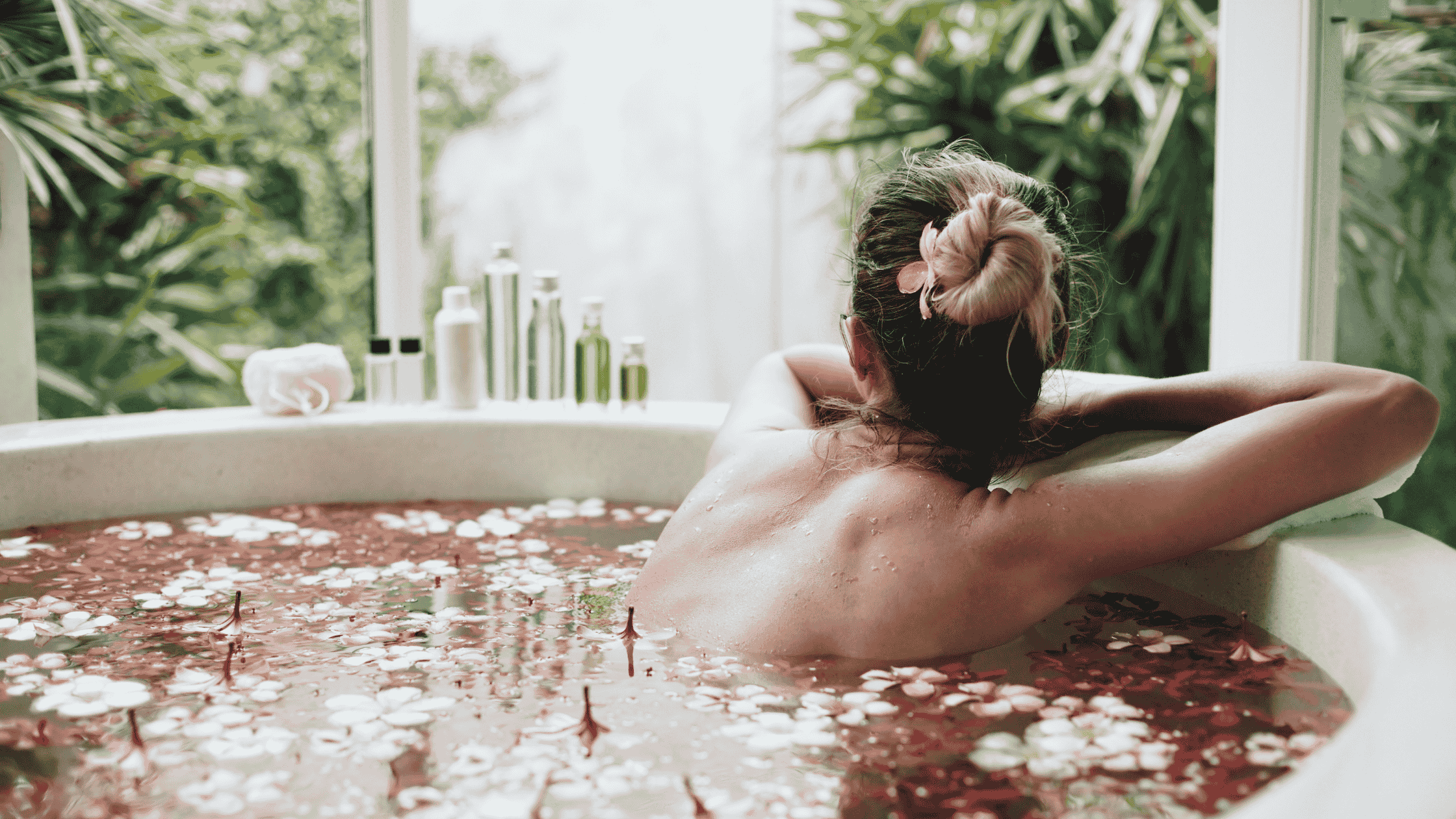 Woman in a bath tub, facing away, head and shoulders uncovered. In a bath of essential oils, and other detox bath ingredients.