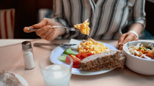 Close up of a plate of food, bread, tomatoes, cucumbers, and a forkful of scrambled eggs. A glass of water is in the foreground. The food is to help lower your blood sugar hormone, insulin.