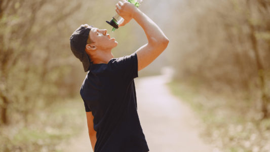 Man wearing a black t-shirt, walking outside. He is drinking form a bottle of water.