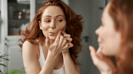 Woman touching her skin blemish in a mirror. She has red hair and a white tank top