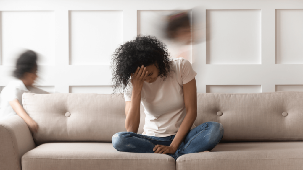 A black woman wearing a white t-shirt and jeans is sitting on a beige couch with her head in her hand. She is sitting cross-legged, and looks exhausted. 2 children, blurred images are running behind her.