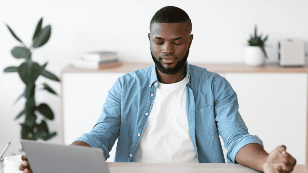 Man with white t-shirt and blue button down, sitting at an office desk learning how to begin meditation. 