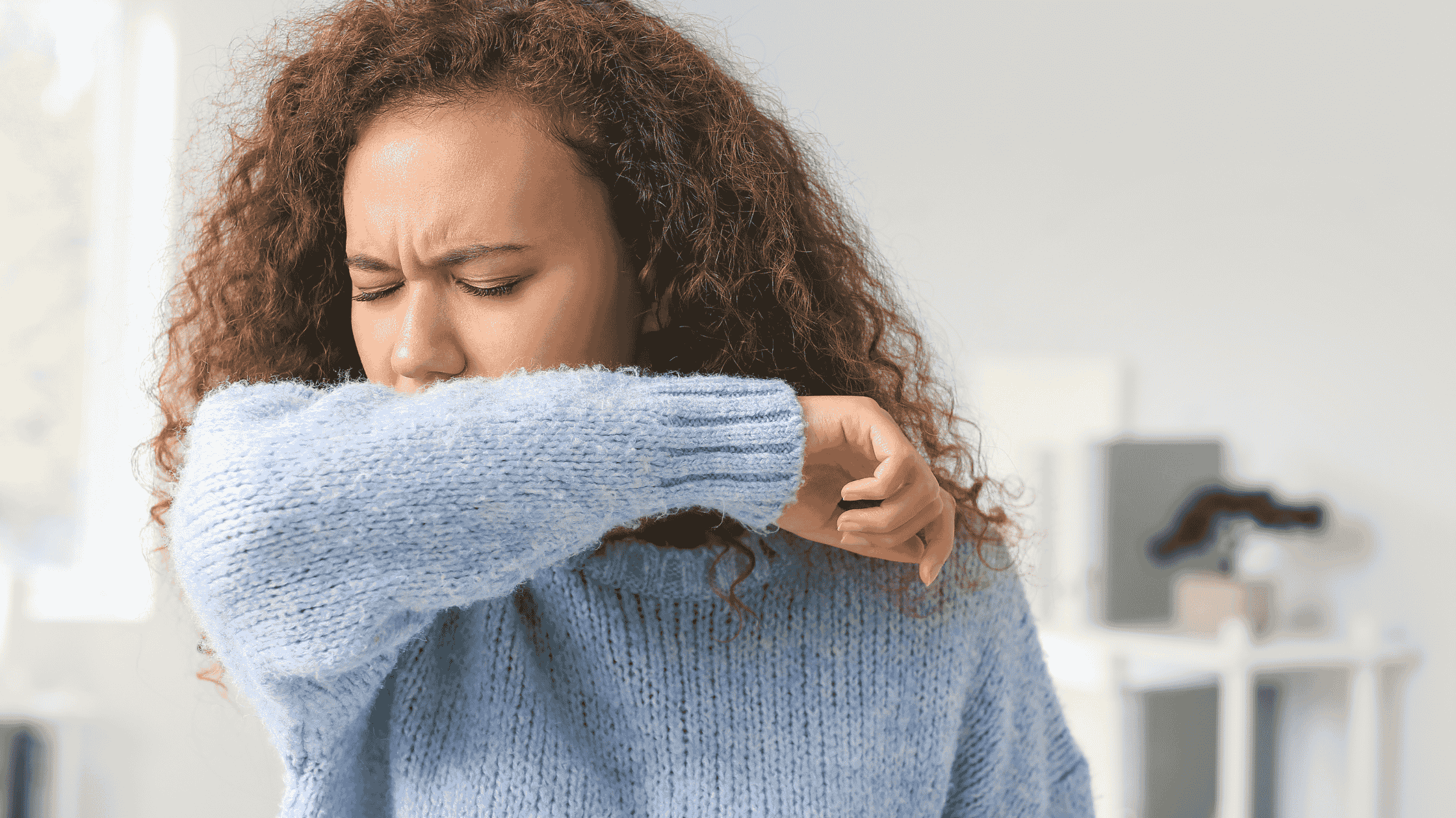 Woman with curly hair, wearing a blue sweater coughing into her arm.