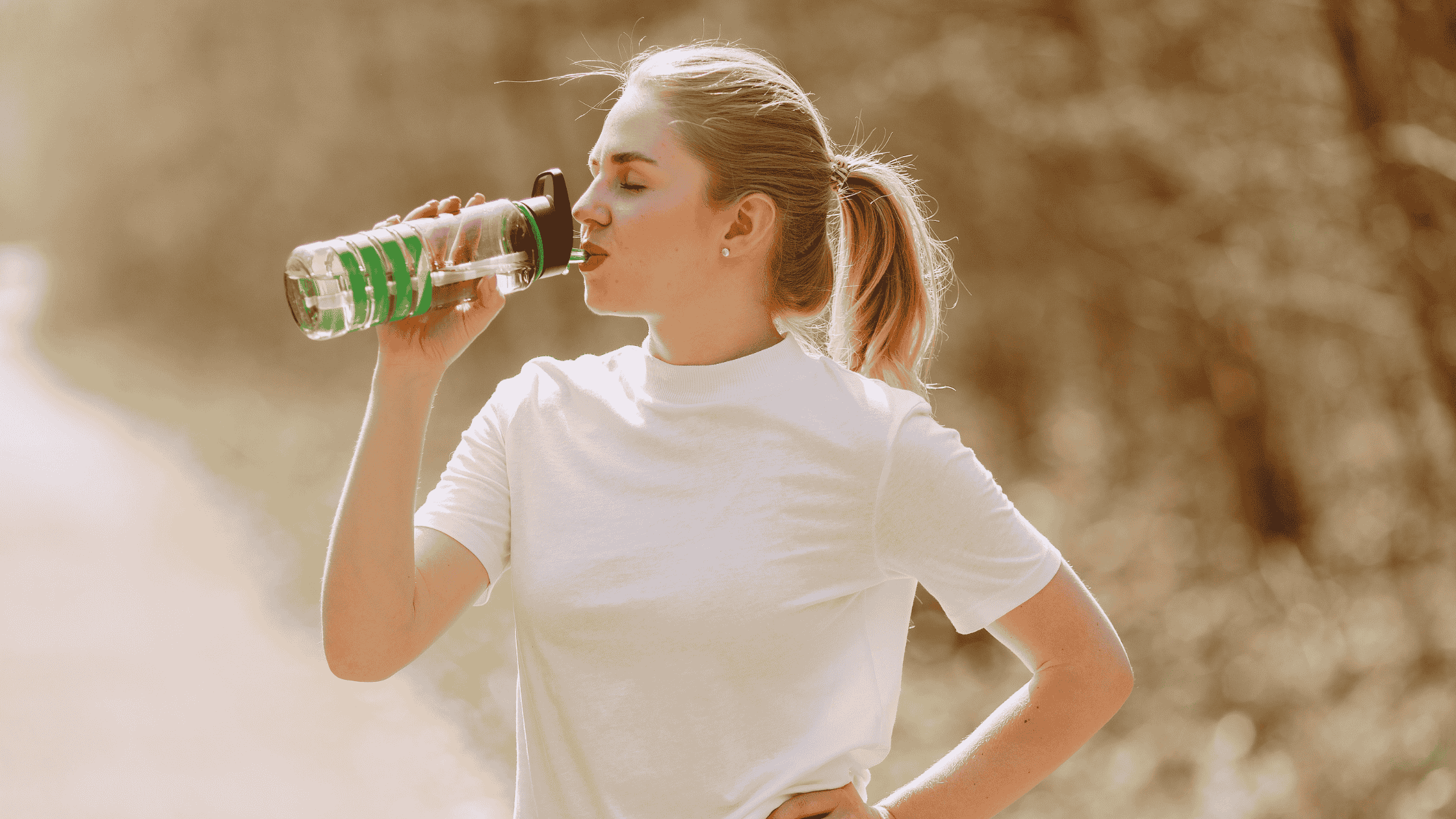 A woman with a blonde pony tail, wearing a white shirt, looks to be walking or hiking. She has stopped to drink water. 