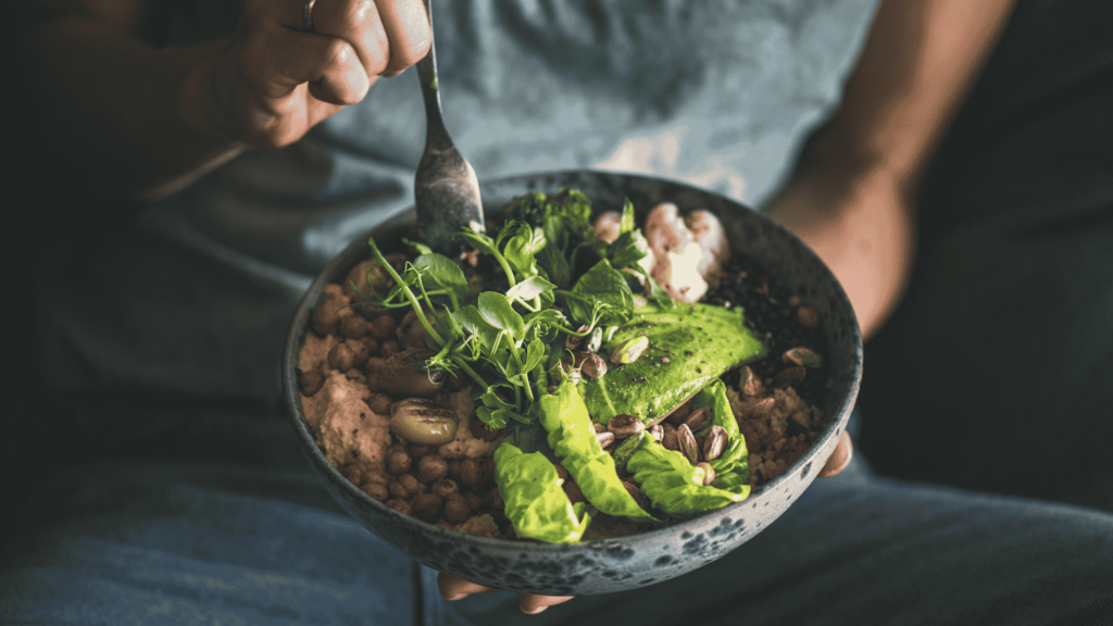 A close up bowl of greens, avocado and other healthy foods. A person is holding the bowl but you only see a fork and a hand.