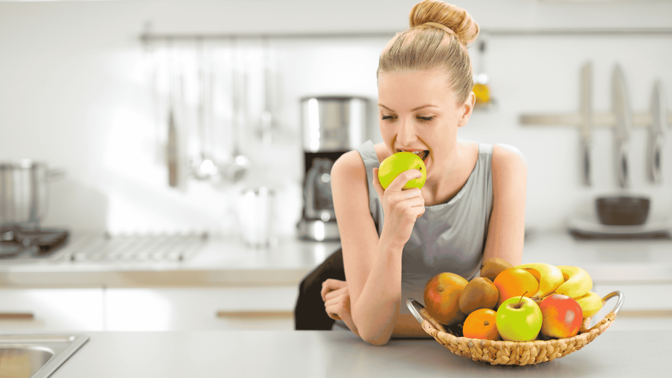 A woman with a high bun and blonde hair, wearing a grey tank top is in a kitchen eating a green apple. 