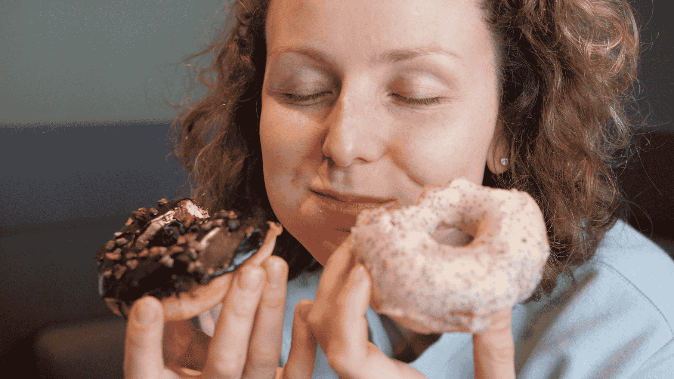 A woman is eating two donuts at the same time. One has a chocolate frosting and one has a pink frosting. This image is used to depict craving carbs and the connection between sugar and candida